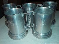 Photo shows 5 metal mugs with brushed finish, each featuring the Playboy bunny logo etched in a square outline on the front. Mugs have angular handles and tiered bases.