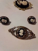 Photo showing a pair of vintage sterling silver filigree cameo earrings, a small vintage sterling silver cameo brooch, and a larger costume cameo brooch with gold-tone frame, all with black backgrounds and white profiles.