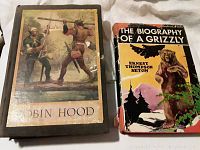 Photo of two vintage books side by side on a fabric surface. Left book is 'Robin Hood' with illustrated cover showing two men crossing a log bridge. Right book is 'The Biography of a Grizzly' with illustrated dust jacket depicting a grizzly bear.