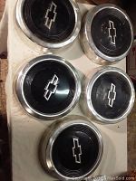 Photo showing five Chevrolet hubcaps arranged on a surface, black centers with silver Chevy logos, chrome edges visible.