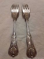 Two silver-tone forks with ornate handles embossed with 'USN' and decorative detailing, placed side by side on a light-colored fabric.