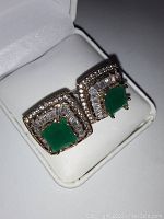 Pair of square-shaped earrings displayed in a white jewelry box showing green synthetic emerald center stones surrounded by rectangular white topaz stones and gold overlay over sterling silver.