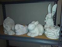 Four rabbit yard art figurines placed on a metal shelf showing all from the side perspective.