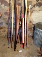 Group of various wooden and metal tool handles and poles leaning against a wall, next to a metal trash can.