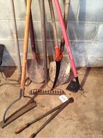 Photo showing a variety of outdoor garden tools including shovels, rakes, hoes, a powered weed cutter, all leaning against a wall indoors.