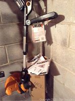 Photo showing the Worx rechargeable string trimmer standing against a concrete wall with manuals and original box at the base.