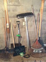 Full view of snow shovels, leaf rakes, metal rakes, and roof snow rake with extension handles leaning against a wall