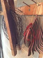 Two wooden frames holding coiled heavy gauge wire and multiple red metal tiller blades with mounting holes hanging beside them.