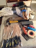 Overview of mixed hardware and tools including screwdrivers, hammer, measuring tape, paint brushes, scrapers, and a grinding stone, some items new in packaging.