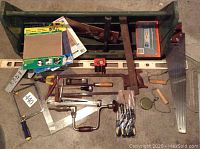 Top-down view of the assortment showing saw blades, sanding sheets, hand crank brace, and various hand tools inside and around the green toolbox.
