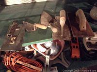 Masonry hand tools mostly plastering and finishing trowels with wooden handles, an orange extension cord, and some other equipment beneath on a green surface.