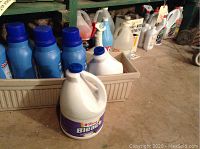 Photo showing several blue jugs of Final Touch laundry detergent and white gallon jugs of Tom's Bleach inside and beside a beige plastic tub on the floor.