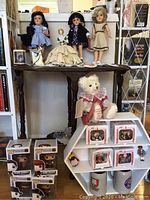 Overall display: three full-size dolls on table top (Toni Dorothy, black-clad, beige dress), tea cozy doll, white bear, small trinket box and pushpins; white shelf below holding boxed figurines and Pop! figures.