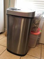 Stainless steel sensor garbage can with black base next to two stacked clear plastic tubs under a window on tile floor, showing overall lot items