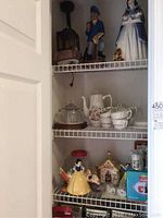Three wire shelving units displaying various items: top shelf lamp, sailor and Snow White figurines; middle shelf glass cheese dome, coffee pot, cups; bottom shelf Snow White cookie jar, ceramic house, s&p sets.