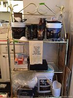 Brass-colored metal bakers rack with popcorn buckets, cups, kitchen towel, and small household items on shelves.