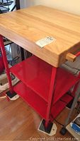 Side view showing butcher block wood top with chips and red metal frame with shelves below.