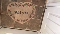 Heart shaped beige welcome mat with floral border and 'Welcome' text placed on tiled floor