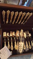 Wooden chest with Rogers Bros silver plated flatware showing eight ornate forks in top row and spoons, servers and carving tools in bottom row