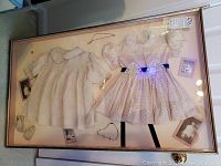 Framed display of two antique children's dresses with accessories and vintage photos.