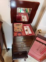 Jewelry armoire with open hinged top showing mirror and jewelry inside; side door open showing necklace hooks and pink lining; drawers below with brass handles.