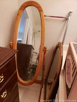 Standing oval mirror with wooden frame and turned posts, showing the length and height with surrounding furniture for scale.