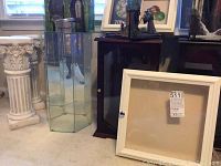 Shows two white ceramic pedestals with classical column detailing, a hexagonal glass display case, dark wood framed glass door display case, and a white framed shadow box display case with a glass front and door knob.