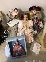 Photo showing five collectors dolls, including porcelain and plastic, several doll stands and original boxes near glass display case.