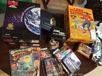 Photo of various boxed dolls and collectibles including G.I. Joe Mercury Astronaut, Star Trek Voyager dolls, The Lone Ranger doll, Star Trek Next Generation box, Superman comic books laid out on a table.