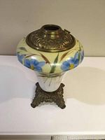 Front view of oil lamp showing the painted glass body with blue flowers and the metal top with embossed decoration.