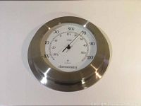 Front view of 2000 Design outdoor thermometer showing round metal frame and white dial with temperature readings.