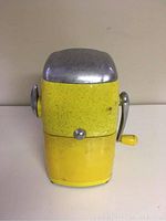 Front view of vintage yellow Ice O Mat ice crusher machine with metal top and hand crank.