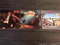 Three vinyl albums by AC/DC laid out on a dark surface: 'High Voltage', 'Let There Be Rock', and 'Dirty Deeds Done Dirt Cheap'.