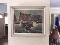 Framed print painting showing boats on the water with cliffs in background, hanging on wall in room.