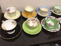Overview of seven assorted bone china tea cups and saucers showing floral designs and different base colors arranged on a black surface.