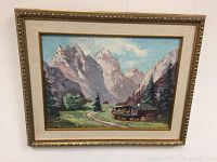 Framed oil painting showing a mountain landscape with a cabin and greenery under a blue sky.