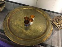 Large round brass plate with detailed etched patterns and four enameled brass shot glasses placed on top. Plate shows some tarnish and wear.