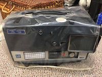 Front and side view of Dejur slide projector with plastic protective cover exploring overall condition and visible brand markings.