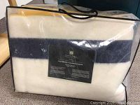 Front view of blanket in clear zippered bag showing folded cream wool with blue stripe and black label
