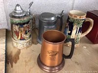 Four beer mugs shown on wooden surface, including two ceramic steins, pewter Molson mug, and copper Niagara Falls mug.