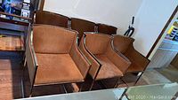 Front and angled view of 6 brown microfiber dining chairs with chrome metal frames arranged in two rows in a room with hardwood flooring.