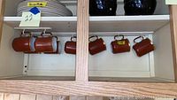 Eight brown ceramic mugs hanging on hooks in a cabinet, shown from above and at an angle, demonstrating their shape, finish, and quantity.
