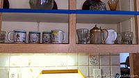 Wood cabinet shelf containing four southwest-themed ceramic mugs, an alabaster mug with metal lid, and clear glass cups
