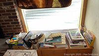 Photo showing art books, sketchbooks, palettes, brushes, and other art supplies arranged on a bench by a window