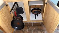 View inside lower cabinet showing assorted frying pans hanging and stacked inside, including black, copper-colored pans and small black pan on shelf.
