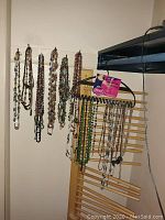 Photo showing multiple necklaces and chains hanging on wall hooks and a wooden rack, displaying a variety of bead styles and metal chains.