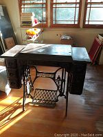 Singer treadle sewing machine with ornate carved wooden drawers and black cast iron treadle base in a room with wooden floor and windows.