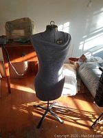 Full front view of grey knit fabric stretch dress form on black tripod metal stand in a room with wooden floor and natural light.