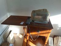 Vintage Singer electric sewing machine with wooden cabinet and drop-leaf extension, dust cover on top, foot pedal placed on cabinet surface.