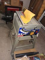 Full view of Ryobi table saw on stand with dust collection attached and yellow adjustment dial visible on top.